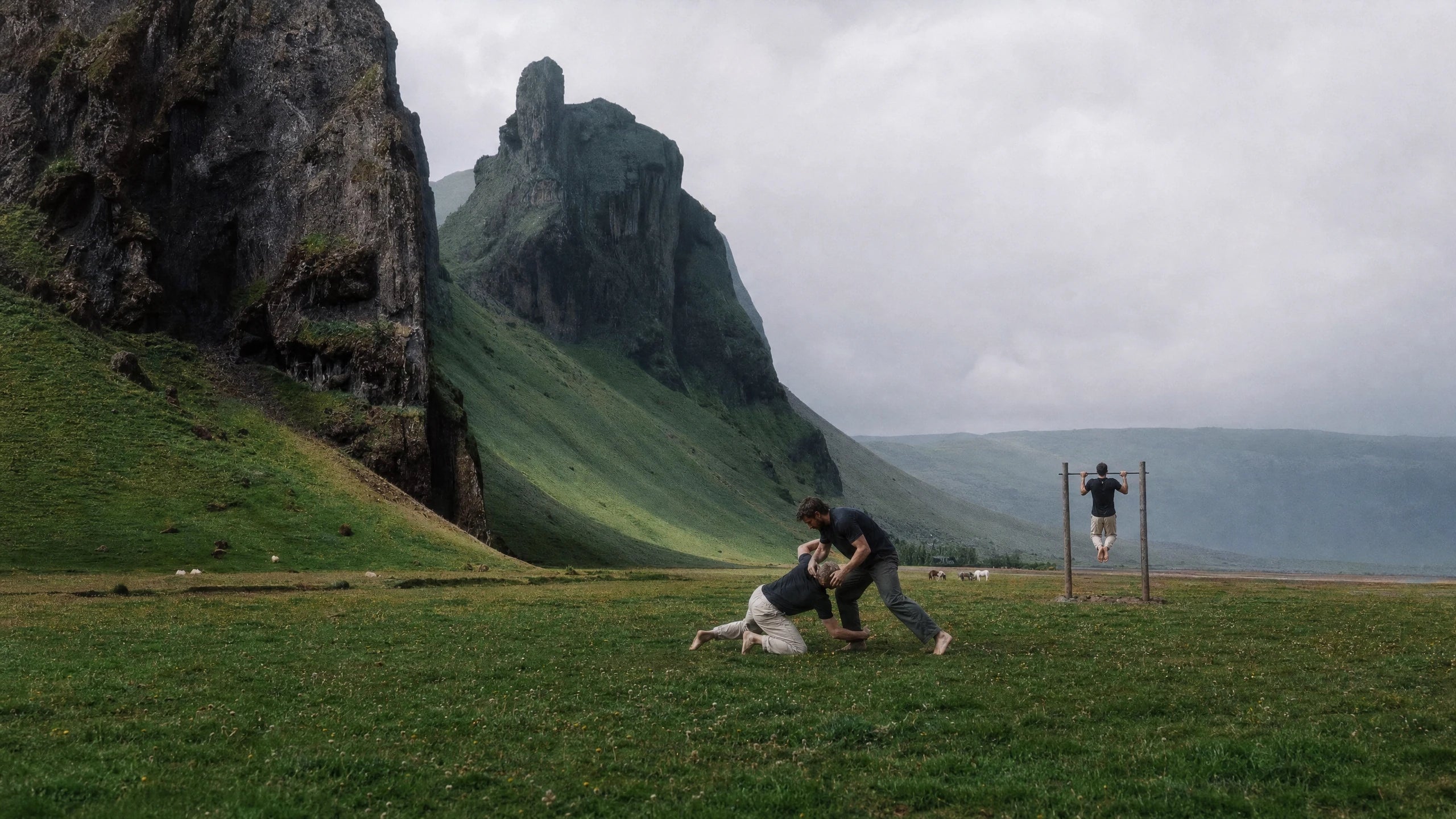 Two people grappling and another doing a pull-up in a grassy landscape beneath tall cliffs.